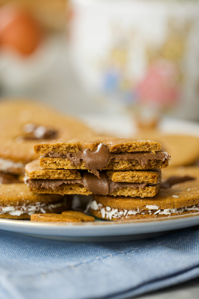 Biscoitos de coco recheado com nutella, empilhado em um prato, com outros biscoitos e detalhes de coco ralado ao fundo.