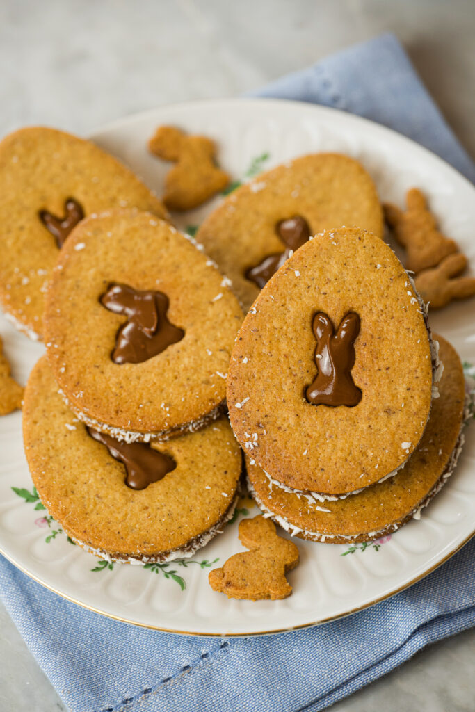 Biscoitos de coco com nutella, de Páscoa em formato oval, com recheio de chocolate em forma de coelho, dispostos em um prato decorado, acompanhados de pequenos biscoitos de coelhinho.
