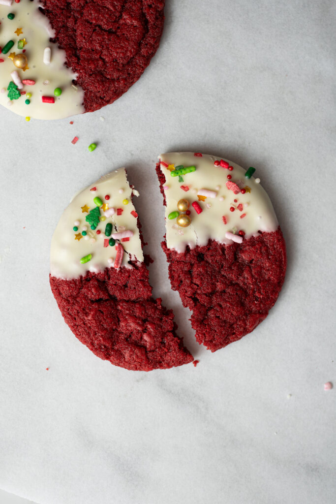 Cookie Red Velvet de Natal com chocolate branco, partido ao meio, mostrando textura macia e decoração com confeitos natalinos.