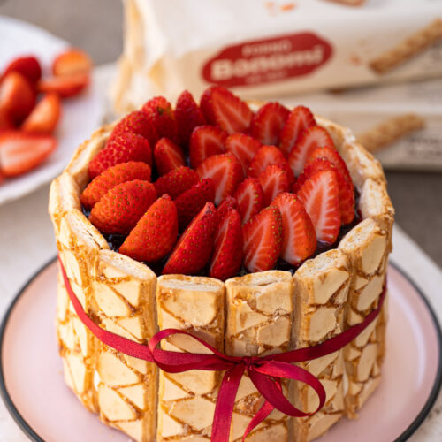 Charlote de chocolate branco com morango inteira vista de cima, decorada com morangos fatiados e laço de fita vermelha, com embalagens da Forno Bonomi ao fundo